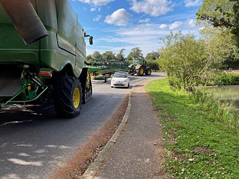 Two large green agricultural vehicles at the road junction by the pond, with a small white car between them.